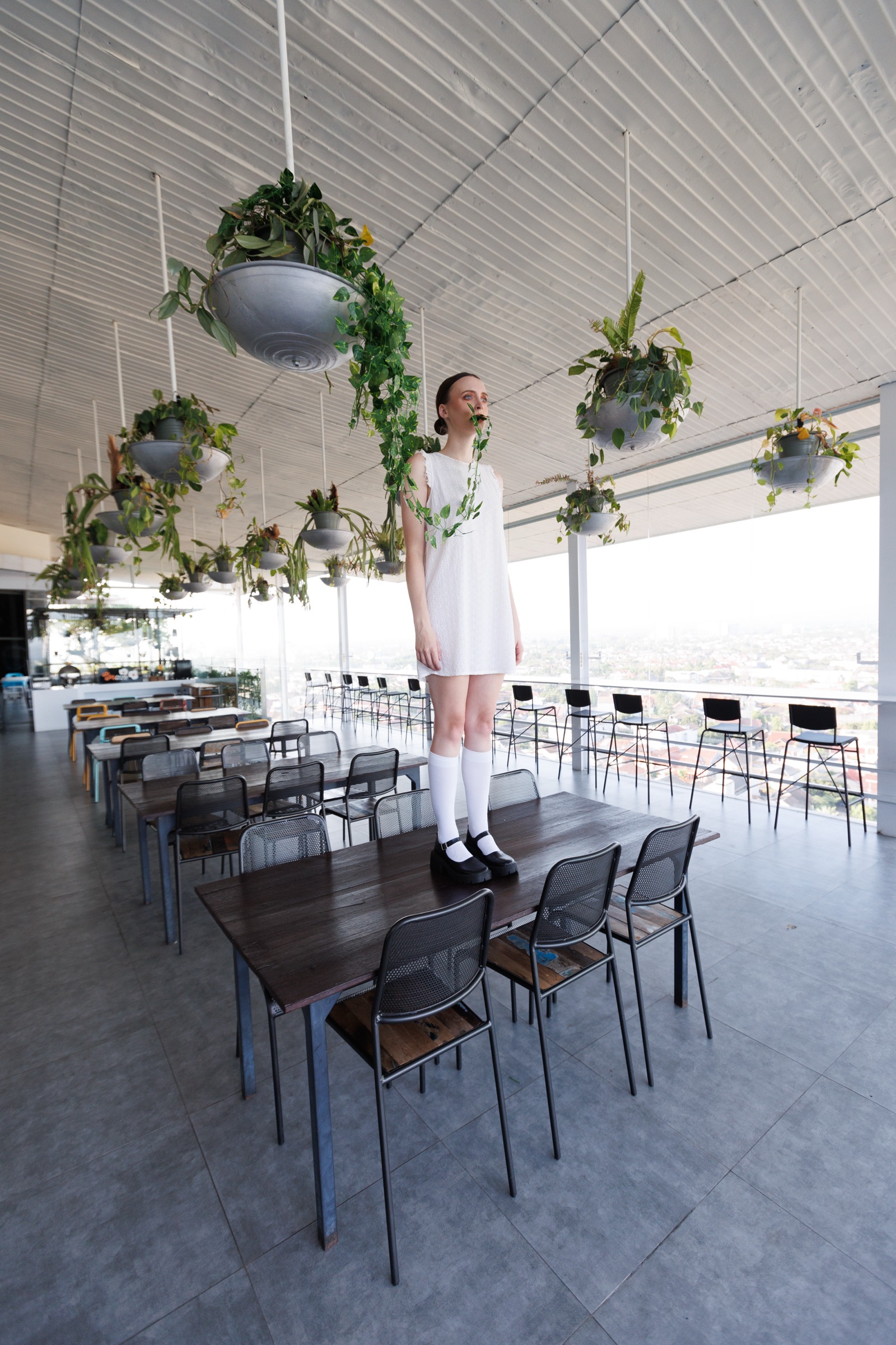 Model in a white dress standing on a table at Alfresco Dining, hanging plants overhead and Jogja beyond